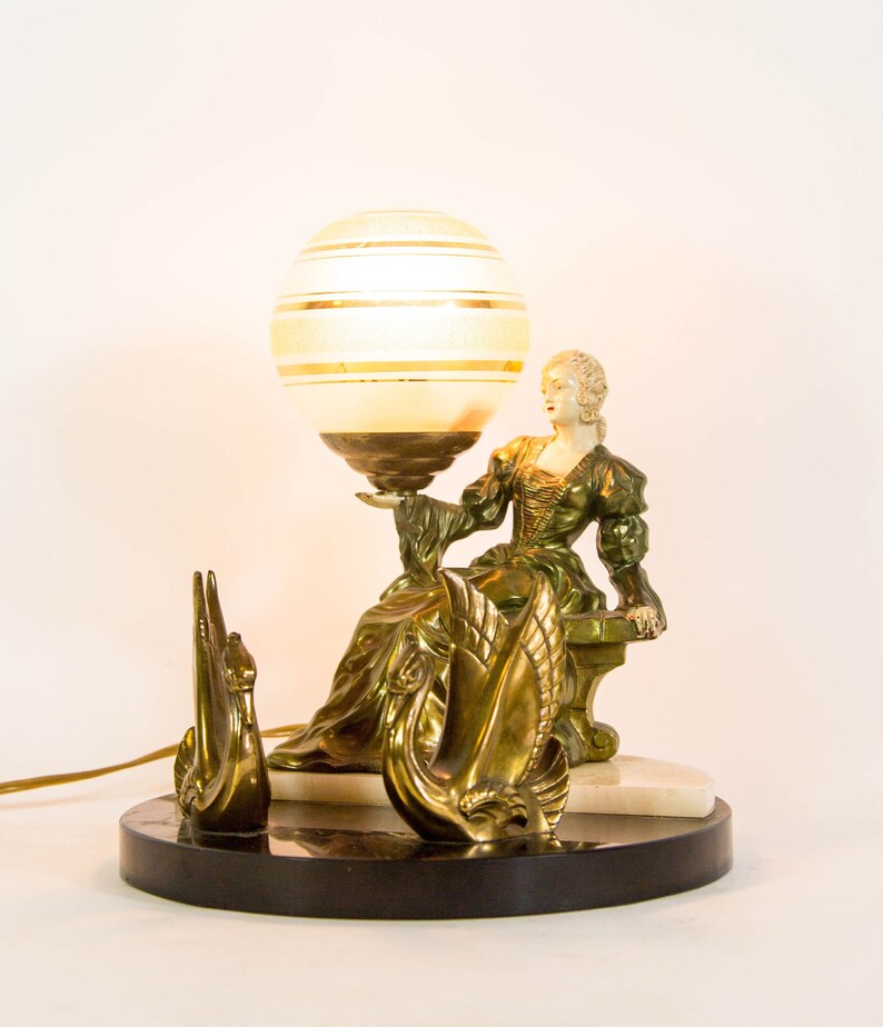 Puede incluir: Una l&aacute;mpara de mesa vintage con acabado dorado presenta a una mujer sosteniendo una pantalla de l&aacute;mpara en forma de globo con rayas doradas. La base de la l&aacute;mpara es de m&aacute;rmol y presenta dos esculturas de cisnes.