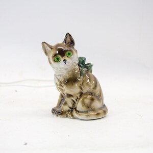 May include: A ceramic figurine of a brown and white cat with green eyes. The cat is sitting with a green bow around its neck.