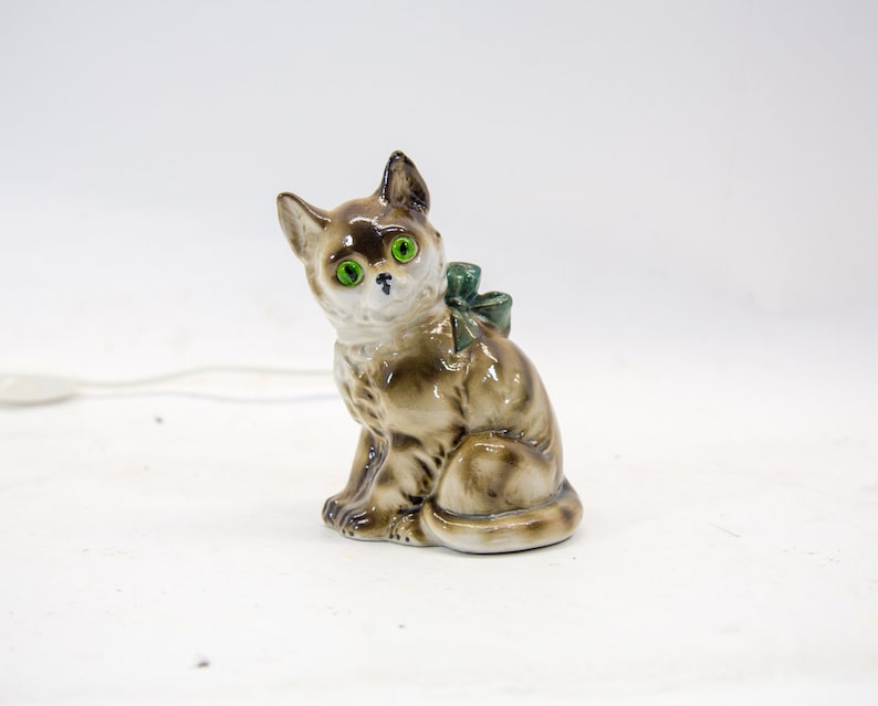 May include: A ceramic figurine of a brown and white cat with green eyes. The cat is sitting with a green bow around its neck.