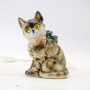 May include: A ceramic cat figurine with green eyes and a green bow. The cat is sitting on a white surface.