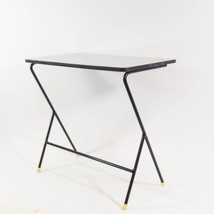 May include: A black metal table with a white top and gold accents. The table has a unique, angled design with a single horizontal support bar.