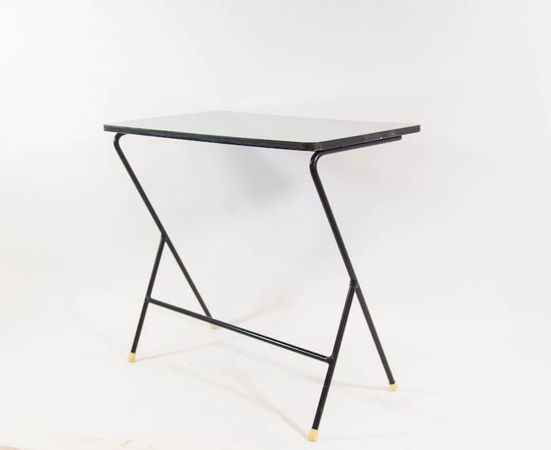 May include: A black metal table with a white top and gold accents. The table has a unique, angled design with a single horizontal support bar.