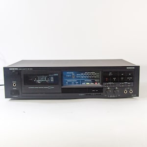 May include: A black Onkyo TA-2570 cassette deck with a digital display and various control knobs. The deck features a cassette tape slot, a headphone jack, and a power button. The unit is on a white background.
