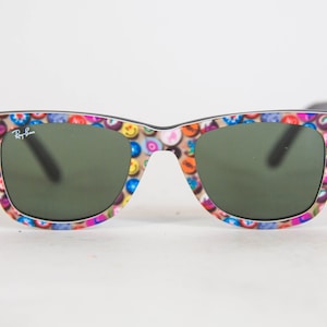 Puede incluir: Gafas de sol Ray-Ban con una montura de patr&oacute;n multicolor de peque&ntilde;os iconos circulares, incluyendo caras sonrientes y varios s&iacute;mbolos, sobre un fondo beige. Las lentes son de color verde oscuro y el logotipo de Ray-Ban aparece en la lente izquierda.