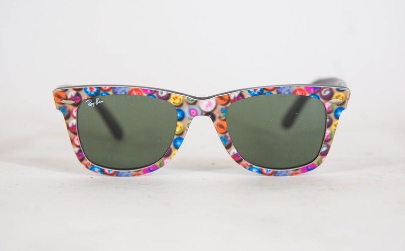 Puede incluir: Gafas de sol Ray-Ban con una montura de patr&oacute;n multicolor de peque&ntilde;os iconos circulares, incluyendo caras sonrientes y varios s&iacute;mbolos, sobre un fondo beige. Las lentes son de color verde oscuro y el logotipo de Ray-Ban aparece en la lente izquierda.