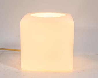 May include: A square, white, illuminated lamp with rounded corners. The lamp emits a soft, warm glow from the open top. A thin, yellow electrical cord is visible on the left side. The lamp is a modern home decor item.