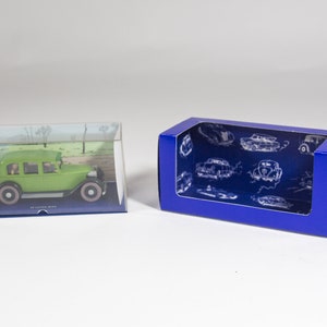 May include: A green toy car in a clear plastic display case with a blue base. The car is a replica of a vintage car with a driver and passengers inside. The case is sitting on a white surface next to a blue cardboard box with a clear window. The box has a pattern of vintage cars on it.