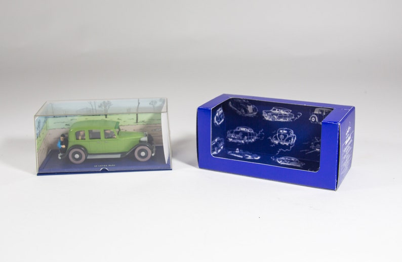 May include: A green toy car in a clear plastic display case with a blue base. The car is a replica of a vintage car with a driver and passengers inside. The case is sitting on a white surface next to a blue cardboard box with a clear window. The box has a pattern of vintage cars on it.