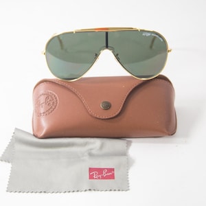 May include: Gold-framed aviator sunglasses with green lenses and a brown leather case. The case has a Ray-Ban logo embossed on the front.
