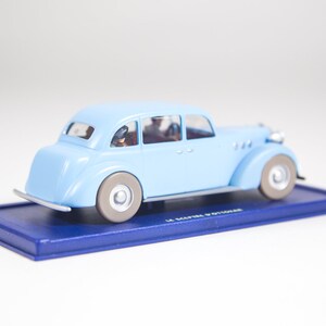 May include: A light blue toy car model with four doors and a black base. The car has a black roof and black wheels. The car is on a blue base with the text "Le Maitre d'Outoir" printed on it.