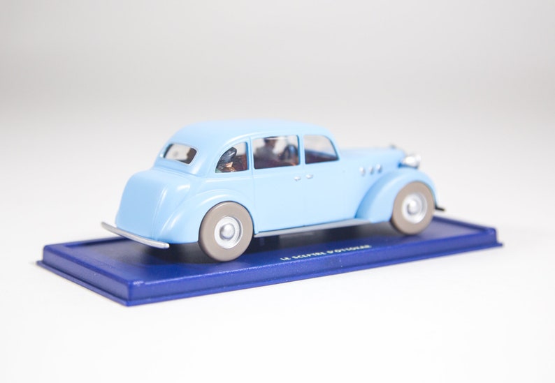 May include: A light blue toy car model with four doors and a black base. The car has a black roof and black wheels. The car is on a blue base with the text "Le Maitre d'Outoir" printed on it.