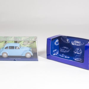 May include: A light blue toy car in a clear plastic box with a green and white background. The car is a 1930s model and is in a blue box with a white background and a picture of a car on it.