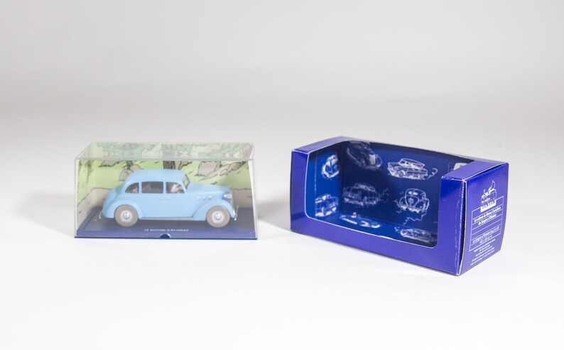 May include: A light blue toy car in a clear plastic box with a green and white background. The car is a 1930s model and is in a blue box with a white background and a picture of a car on it.