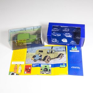 May include: A green toy car in a box with a comic book featuring Tintin and the Blue Lotus. The car is a replica of the one driven by Tintin in the comic book.