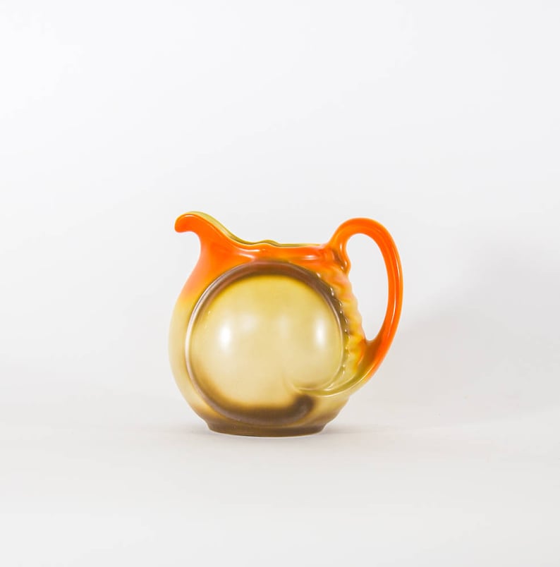 May include: A ceramic pitcher with a brown and orange glaze. The pitcher has a rounded body and a handle.
