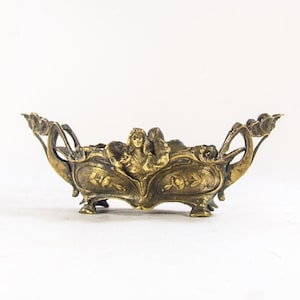 Op de afbeelding: Oud messing kom met gedetailleerd ontwerp. De kom heeft gebeeldhouwde figuren, bloempatronen en gebogen handvatten. Hij staat op kleine voetjes en heeft een ondiep, open interieur.