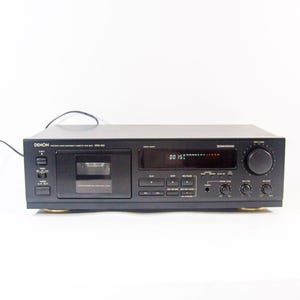 May include: Black Denon DRM-655 cassette deck with a digital display showing "00 15". The deck has various buttons, knobs, and a cassette tape compartment. The unit is on a white surface.
