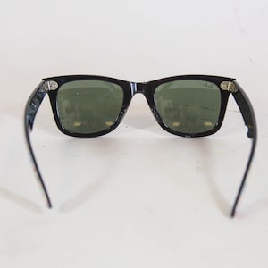 Puede incluir: Gafas de sol negras con una montura rectangular cl&aacute;sica y lentes tintadas en verde oscuro. La montura presenta detalles met&aacute;licos plateados en las bisagras y el logotipo de Ray-Ban est&aacute; impreso en blanco en la lente derecha.