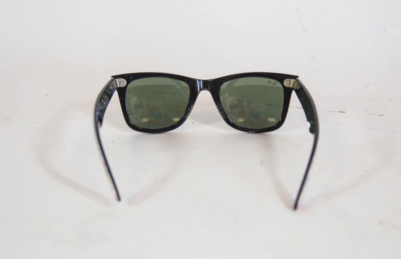 Puede incluir: Gafas de sol negras con una montura rectangular cl&aacute;sica y lentes tintadas en verde oscuro. La montura presenta detalles met&aacute;licos plateados en las bisagras y el logotipo de Ray-Ban est&aacute; impreso en blanco en la lente derecha.