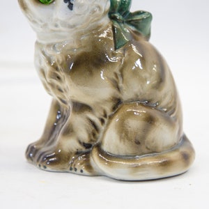 May include: A ceramic figurine of a brown and white cat with green eyes. The cat is sitting with its tail curled around its legs and has a green bow on its back.
