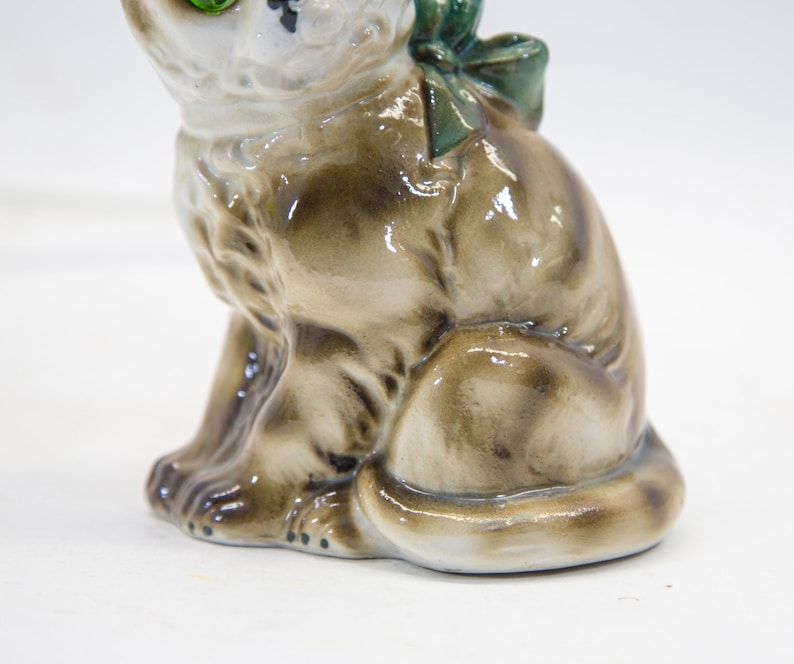 May include: A ceramic figurine of a brown and white cat with green eyes. The cat is sitting with its tail curled around its legs and has a green bow on its back.