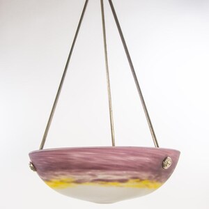 Pu&ograve; includere: Un lampadario a sospensione con un paralume a forma di ciotola. Il paralume presenta una miscela di colori, tra cui viola, giallo e bianco. Il lampadario &egrave; sospeso da tre aste metalliche attaccate a un supporto decorativo a soffitto.