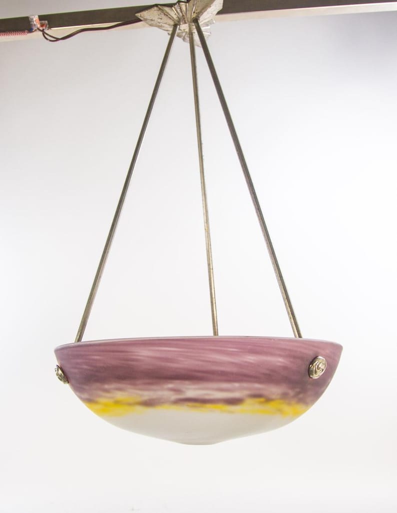 Pu&ograve; includere: Un lampadario a sospensione con un paralume a forma di ciotola. Il paralume presenta una miscela di colori, tra cui viola, giallo e bianco. Il lampadario &egrave; sospeso da tre aste metalliche attaccate a un supporto decorativo a soffitto.