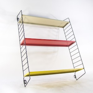 May include: A three-tiered wall shelf unit with cream, red, and yellow shelves. The frame is black metal. The shelves are rectangular. The unit is designed to be mounted on a wall, and is likely made of metal or wood.