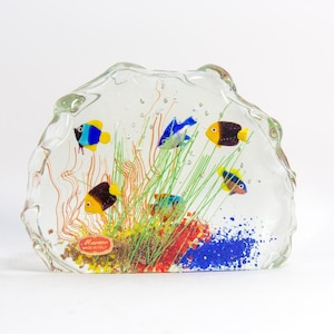 May include: A clear glass sculpture with colorful fish and aquatic plants. The sculpture features yellow, blue, and black fish swimming among green and red plants. The base has a blue and red speckled design. The label reads "Murano Made in Italy."