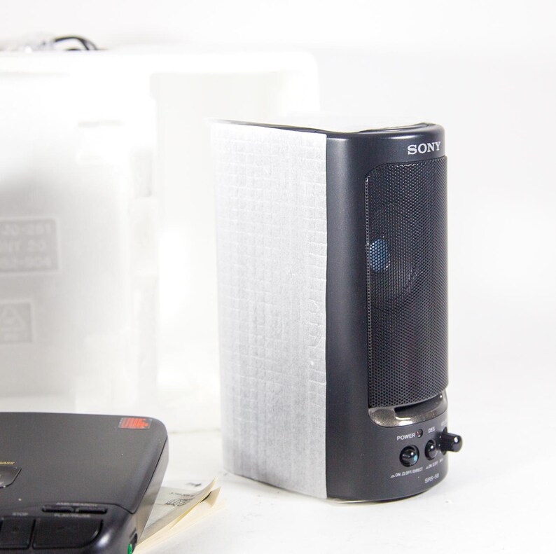 May include: A black Sony speaker with a silver trim and a black speaker grille. The speaker has a power button, volume control, and a headphone jack. The speaker is sitting on a white surface.