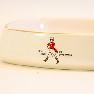 May include: White ceramic ashtray with a gold rim. The ashtray features a red and black illustration of a man walking with a cane, and the text "Born 1820 still going strong."