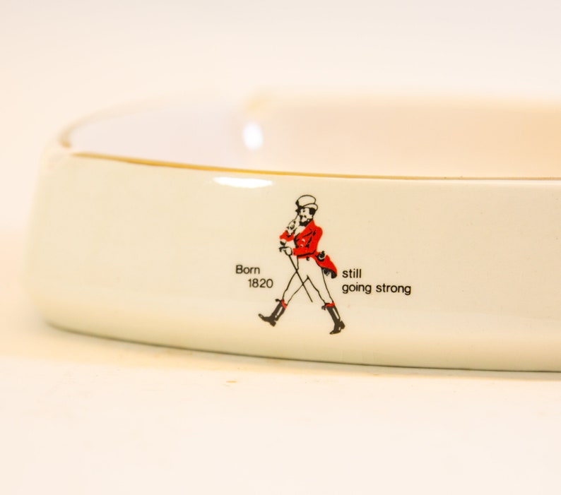 May include: White ceramic ashtray with a gold rim. The ashtray features a red and black illustration of a man walking with a cane, and the text "Born 1820 still going strong."
