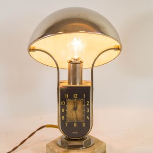Peut inclure: Une lampe de table argent&eacute;e et noire avec un cadran d'horloge et le mot "norma" sur la base. La lampe a un abat-jour en forme de d&ocirc;me et une base en marbre blanc.