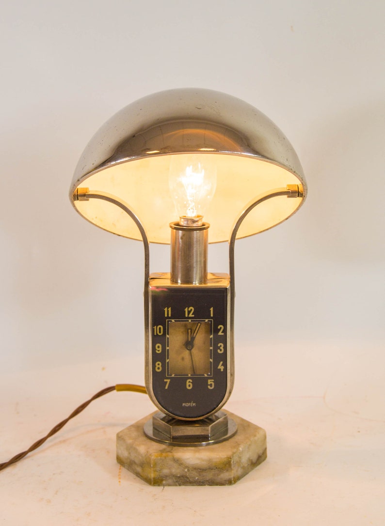 Peut inclure: Une lampe de table argent&eacute;e et noire avec un cadran d'horloge et le mot "norma" sur la base. La lampe a un abat-jour en forme de d&ocirc;me et une base en marbre blanc.