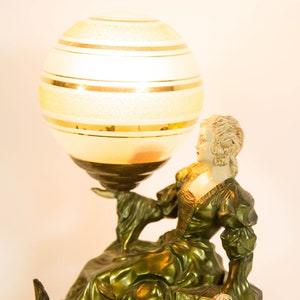 Puede incluir: Una l&aacute;mpara de mesa vintage con una pantalla de globo dorada y blanca y una escultura de bronce de una mujer sosteniendo el globo. La mujer lleva un vestido verde y tiene la cara blanca.