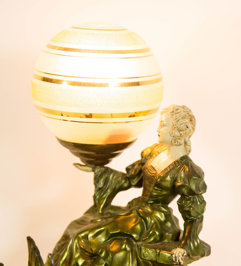 Puede incluir: Una l&aacute;mpara de mesa vintage con una pantalla de globo dorada y blanca y una escultura de bronce de una mujer sosteniendo el globo. La mujer lleva un vestido verde y tiene la cara blanca.