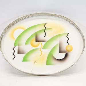 Könnte beinhalten: Ein weißes ovales Serviertablett mit silbernem Rand. Das Tablett zeigt ein geometrisches Design mit grünen, gelben und schwarzen Formen.