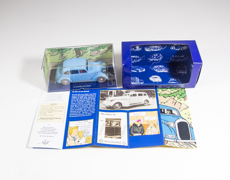 May include: A blue toy car in a clear plastic box with a blue and white cardboard box. The box has a picture of the car and the text "Le Voltigeur", "De auto van het Suskewiet", and "From albums 1953".