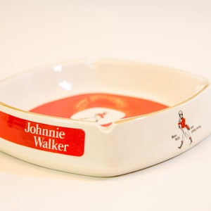 May include: White ceramic ashtray with a red and white interior. The ashtray features the "Johnnie Walker" logo in red and white, along with a graphic of a man in a red coat. The ashtray is a square shape with rounded corners.