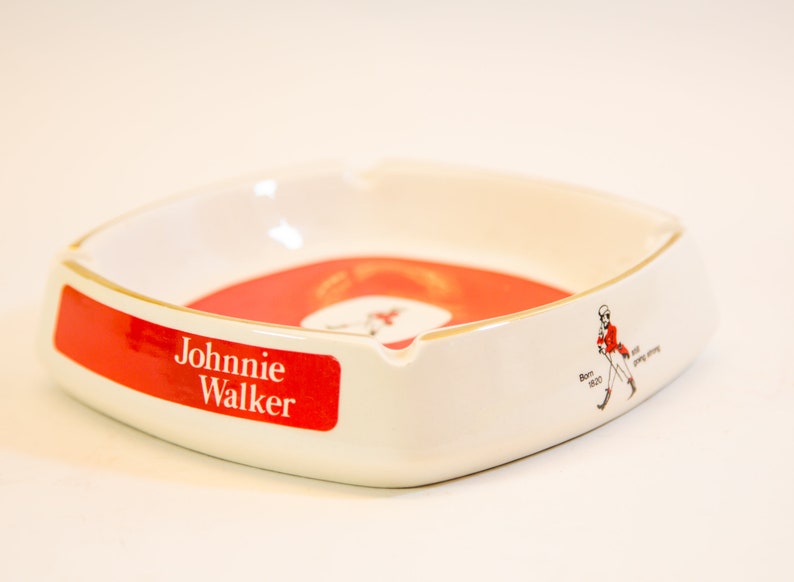 May include: White ceramic ashtray with a red and white interior. The ashtray features the "Johnnie Walker" logo in red and white, along with a graphic of a man in a red coat. The ashtray is a square shape with rounded corners.