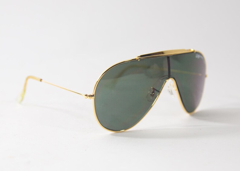 May include: Gold-framed aviator sunglasses with green lenses. The sunglasses have a distinctive, wraparound design.
