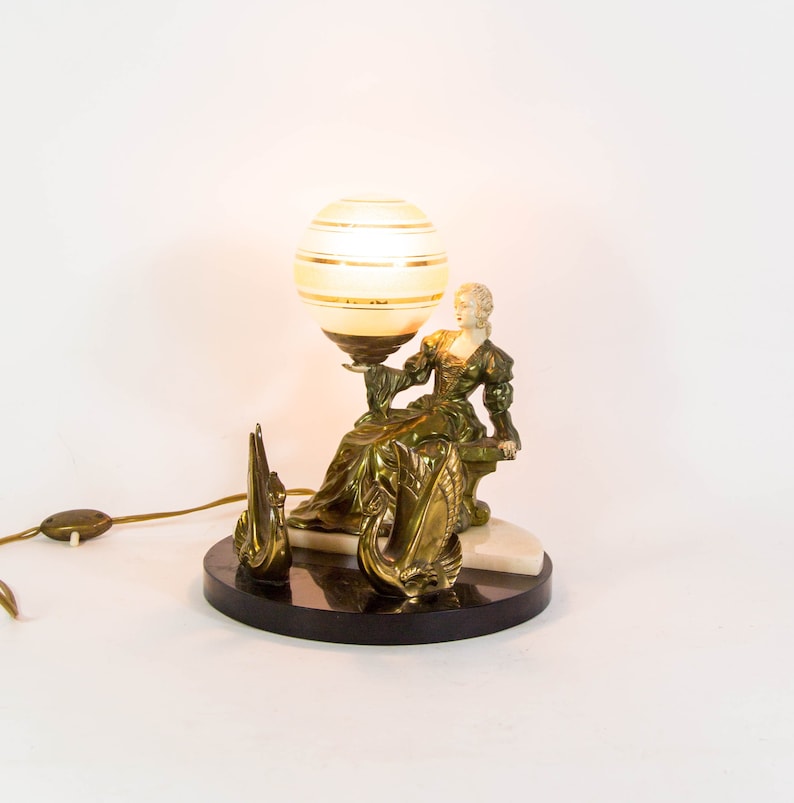 Puede incluir: Una l&aacute;mpara de mesa con acabado dorado presenta una figura de mujer sosteniendo una pantalla de l&aacute;mpara en forma de globo con rayas blancas y doradas. La base de la l&aacute;mpara est&aacute; hecha de m&aacute;rmol negro con un acento de m&aacute;rmol blanco. Dos figuras de cisnes se encuentran a cada lado de la figura de la mujer.