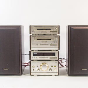 Könnte beinhalten: Ein Vintage-Stereosystem mit zwei dunkelbraunen Lautsprechern und einem Stapel silberner Komponenten. Zu den Komponenten gehören ein CD-Player, ein Tuner und ein Verstärker. Die Lautsprecher sind rechteckig und tragen den Markennamen "Technics".