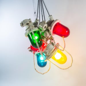 Op de afbeelding: Een cluster van kleurrijke hanglampen in industriële stijl. Elke lamp heeft een heldere, afgeronde glazen behuizing in rood, groen, blauw en geel, met een zilverkleurige metalen armatuur. De lampen hangen aan dunne draden.