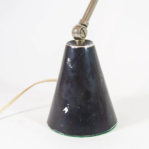 Może przedstawiać: Czarna metalowa lampa biurkowa z kloszem w kształcie stożka i regulowanym srebrnym ramieniem. Lampa ma zieloną filcową podstawę.
