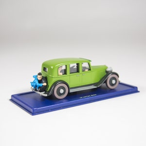 May include: A green toy car with a blue figure sitting on the back bumper. The car is on a blue base with the text "Le Lotus Bleu" printed on it.