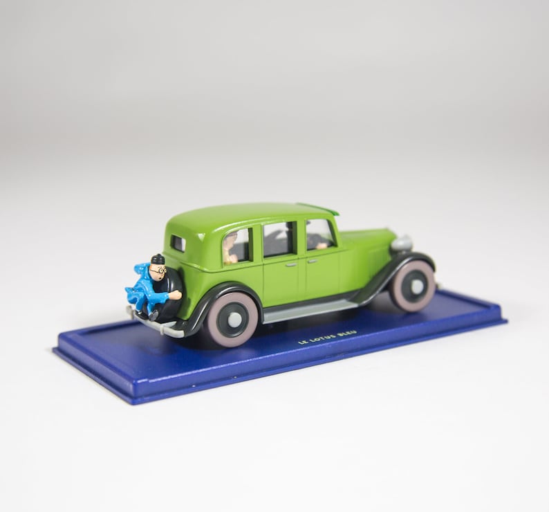 May include: A green toy car with a blue figure sitting on the back bumper. The car is on a blue base with the text "Le Lotus Bleu" printed on it.