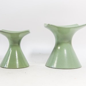 May include: Two light green ceramic pedestal vases with a flared top and a conical base. The vases have a glossy finish and are displayed against a white background. The vases are of different sizes.