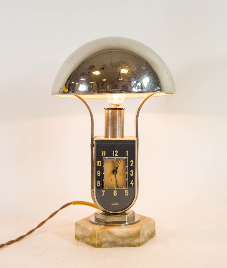 Peut inclure: Une lampe de table argent&eacute;e et noire avec un abat-jour en d&ocirc;me chrom&eacute; et un cadran d'horloge avec des chiffres romains. La lampe est sur un socle en marbre et a un cordon.
