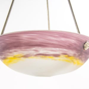 Pu&ograve; includere: Un lampadario in vetro con un paralume a forma di ciotola. Il paralume presenta una miscela di colori, tra cui viola, giallo e bianco. Il lampadario &egrave; sospeso da sottili fili metallici e presenta accenti metallici decorativi.